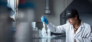 Man wearing a white lab coat, glasses, a black baseball cap, and blue gloves is dispensing liquid from a pipette 