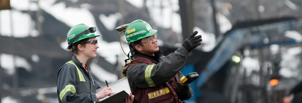Two workers in safety gear at an industrial site, one pointing while the other takes notes.