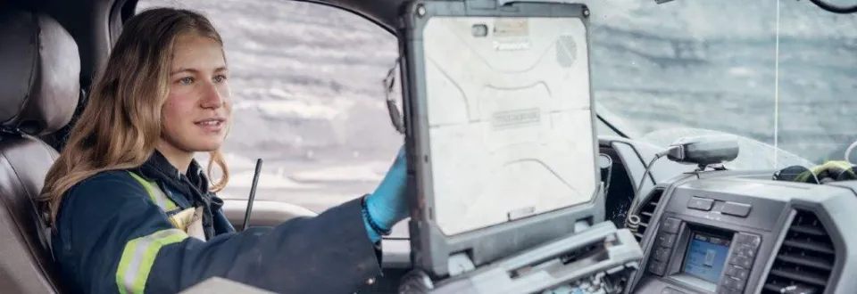 Woman in safety gear using a laptop inside a vehicle at a mining site.